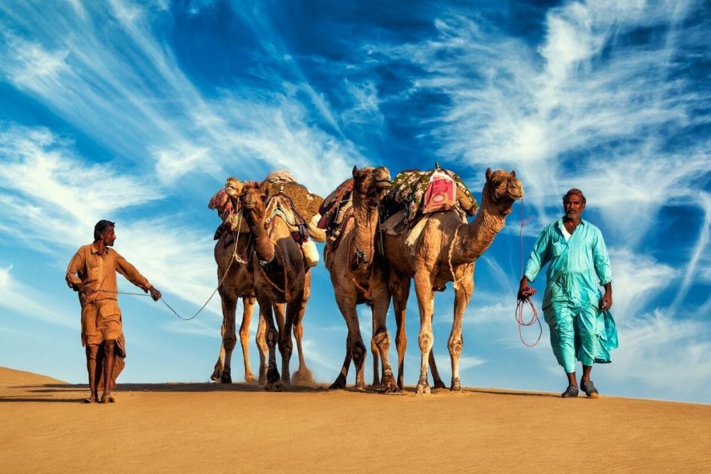 Two camels and two men in the desert with an incredible sky above them