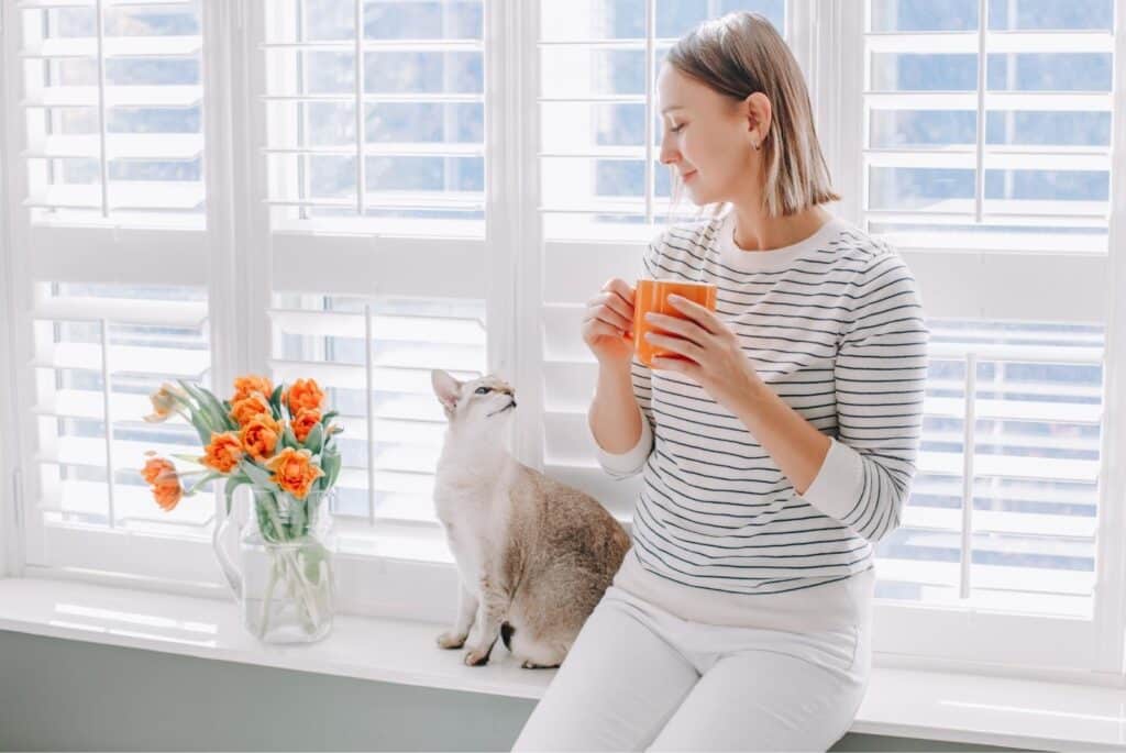 Woman sitting by her window drinking key with her cat - annakraynova - Canva Pro